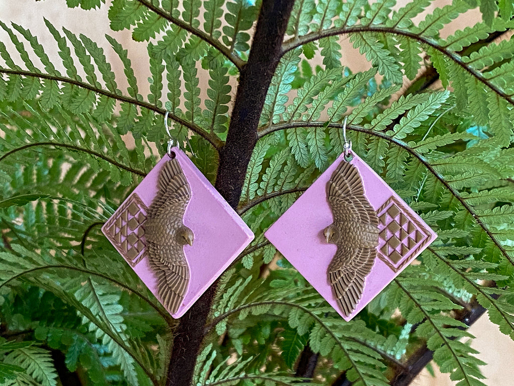 The earrings in a lilac and gold colourway, hanging from a fern frond